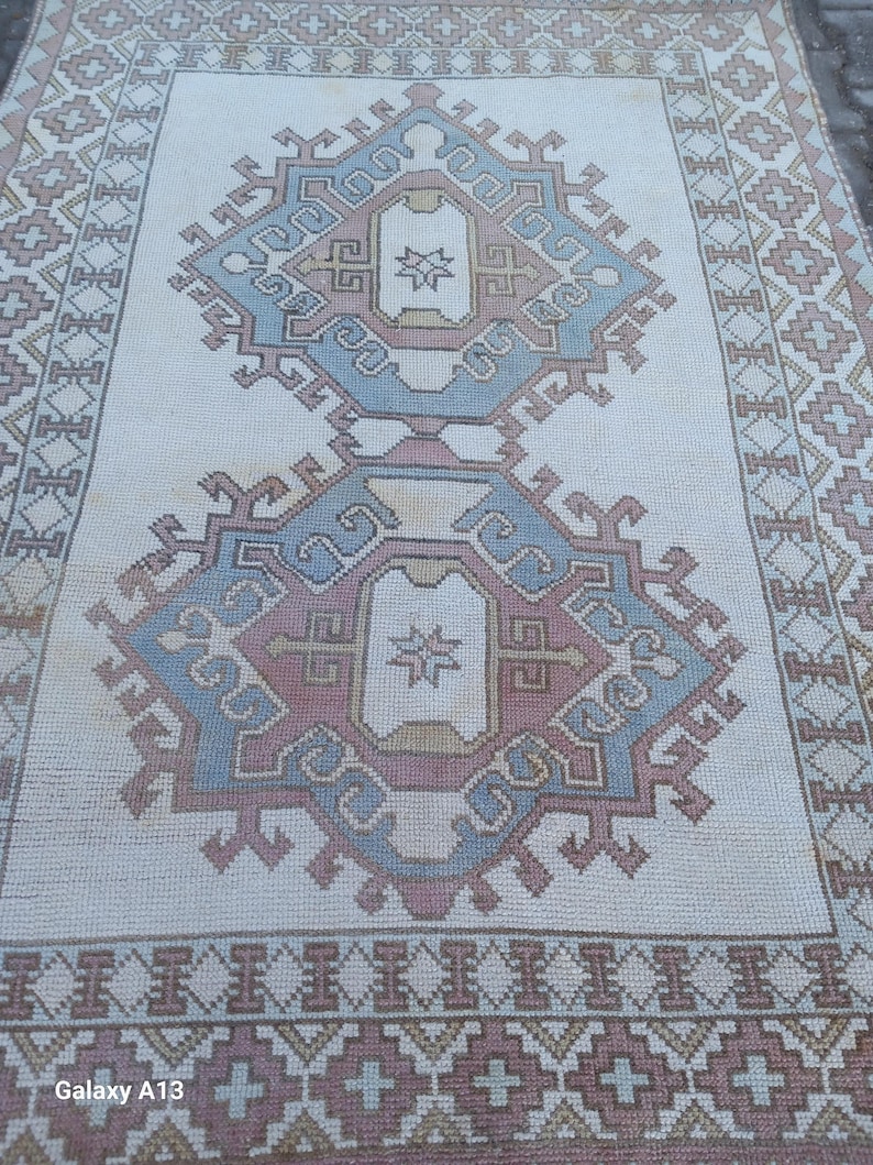May include: A white rug with a blue, pink, and beige geometric pattern. The rug has a border with a repeating pattern of small squares and diamonds.