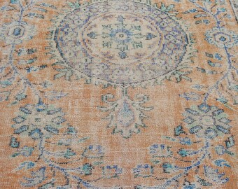 Puede incluir: Una alfombra vintage con un fondo naranja descolorido y un patrón floral azul y blanco. La alfombra tiene un medallón central con un diseño geométrico.