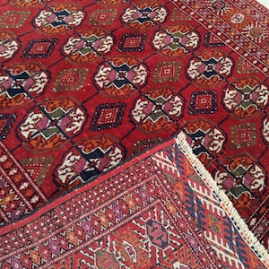 May include: A red oriental rug featuring a repeating geometric design in orange, blue, and white. The rug has a detailed border and a fringed edge, demonstrating traditional craftsmanship.