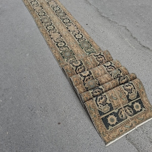May include: A long, narrow runner rug featuring a repeating geometric pattern in shades of black, tan, and muted green. The design includes intricate floral motifs and bordered edges, with the end of the rug folded over itself in several layers.