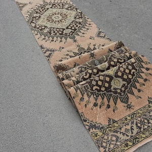 May include: A long, narrow runner rug featuring a faded peach background with repeating dark brown and muted green geometric medallion patterns. The rug is partially folded at one end, revealing the woven texture and the intricate, symmetrical design.