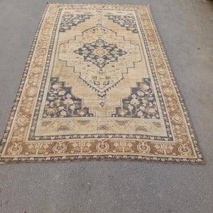 May include: A rectangular beige rug with a central navy blue medallion design and a border of brown and navy blue floral patterns. The rug is placed on a gray surface.
