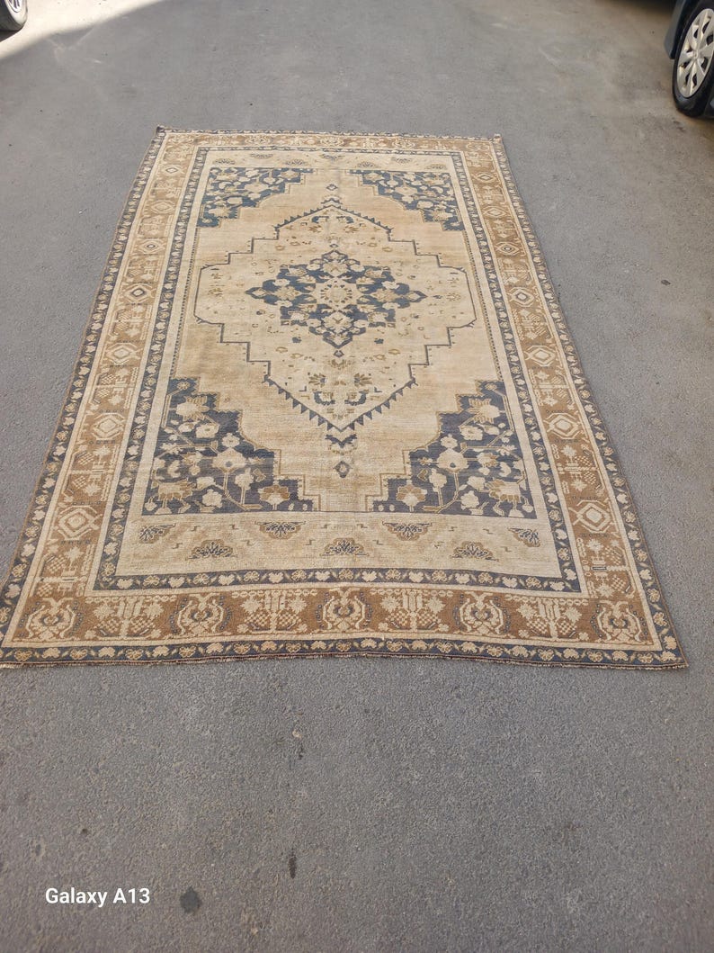 May include: A rectangular beige rug with a central navy blue medallion design and a border of brown and navy blue floral patterns. The rug is placed on a gray surface.