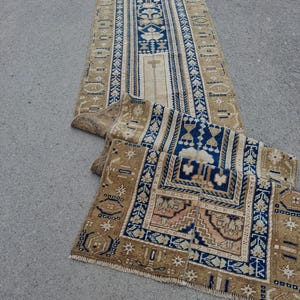 May include: A long, rectangular runner rug featuring a central blue panel with floral motifs, surrounded by beige borders with geometric patterns. The rug displays intricate, symmetrical designs in shades of blue, tan, and cream, with a fringed edge.