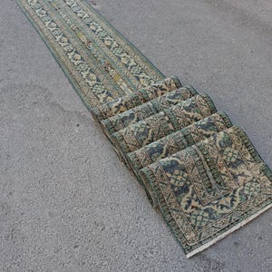 May include: A long, narrow runner rug featuring a repeating geometric and floral pattern in shades of muted teal, sage green, and cream. The rug is partially folded over itself in several layers, displaying a fringed edge at the bottom end.