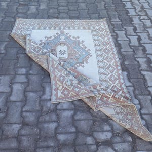 May include: A white rug with a geometric pattern in shades of pink, blue, and beige. The rug is folded in half and lying on a gray concrete surface.