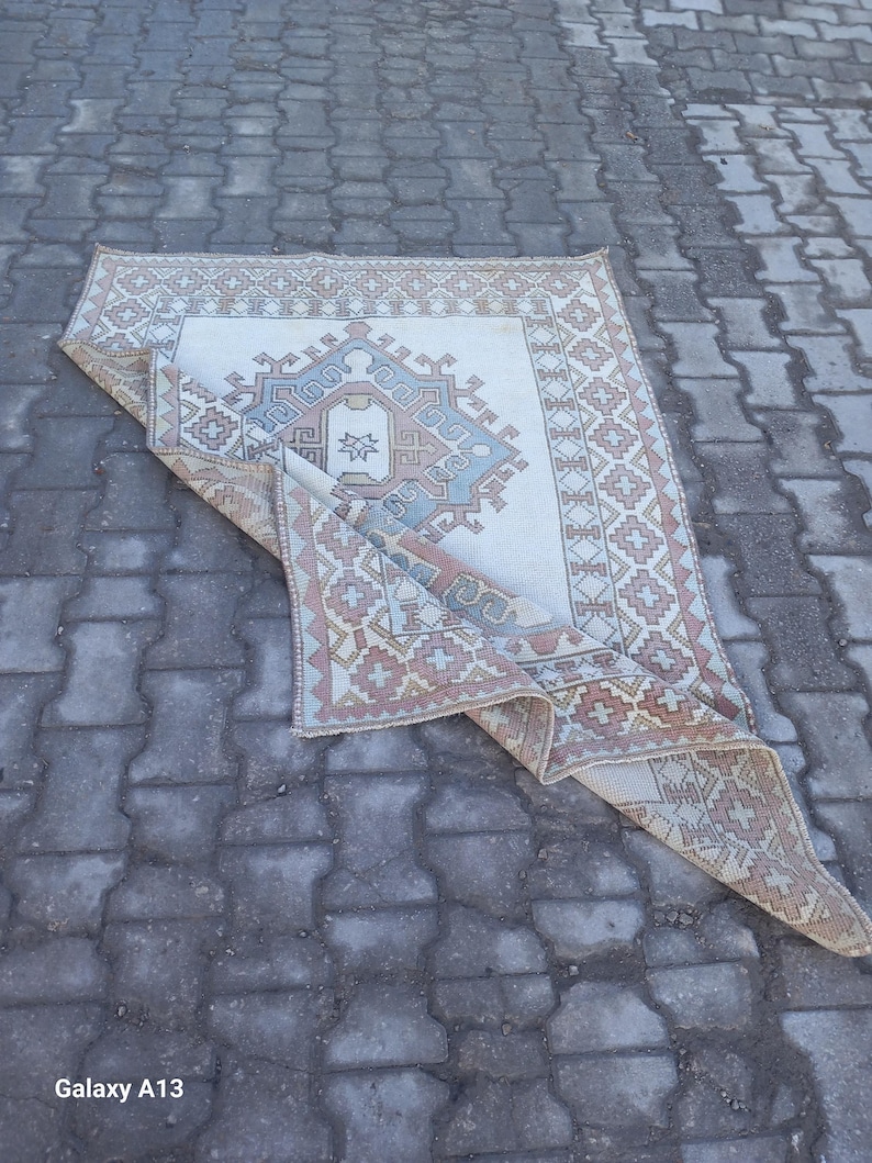 May include: A white rug with a geometric pattern in shades of pink, blue, and beige. The rug is folded in half and lying on a gray concrete surface.