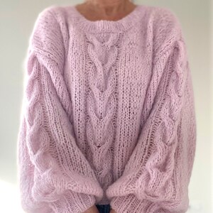 Puede incluir: Un su&eacute;ter rosa claro de gran tama&ntilde;o con cuello redondo y detalles de punto trenzado. Las mangas largas tambi&eacute;n presentan un patr&oacute;n de punto trenzado. El su&eacute;ter est&aacute; hecho de un material suave y texturizado, ideal para climas m&aacute;s frescos.