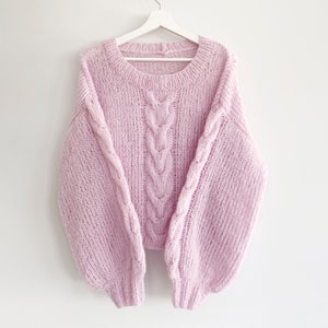 Puede incluir: Un su&eacute;ter rosa claro tejido a mano con un dise&ntilde;o de punto trenzado en la parte delantera y en las mangas. El su&eacute;ter tiene un cuello redondo y mangas largas y voluminosas. Cuelga de una percha blanca sobre un fondo blanco.