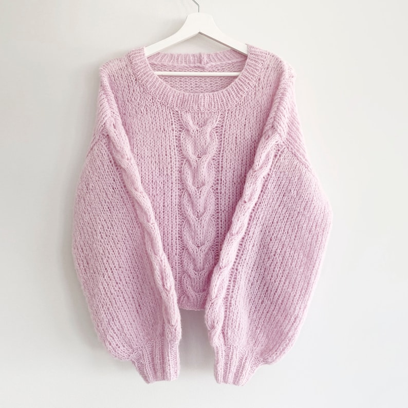 Puede incluir: Un su&eacute;ter rosa claro tejido a mano con un dise&ntilde;o de punto trenzado en la parte delantera y en las mangas. El su&eacute;ter tiene un cuello redondo y mangas largas y voluminosas. Cuelga de una percha blanca sobre un fondo blanco.
