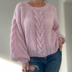 Puede incluir: Un su&eacute;ter de punto rosa claro, tejido a mano, con cuello redondo y detalles de punto trenzado en el centro y en las mangas. El su&eacute;ter tiene un corte holgado y c&oacute;modo, combinado con vaqueros azules.
