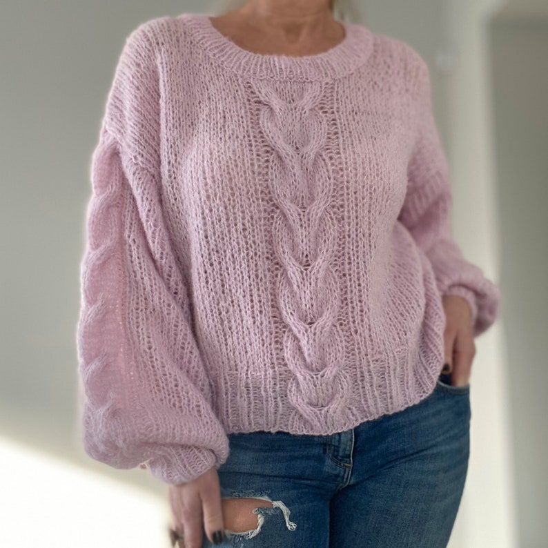 Puede incluir: Un su&eacute;ter de punto rosa claro, tejido a mano, con cuello redondo y detalles de punto trenzado en el centro y en las mangas. El su&eacute;ter tiene un corte holgado y c&oacute;modo, combinado con vaqueros azules.