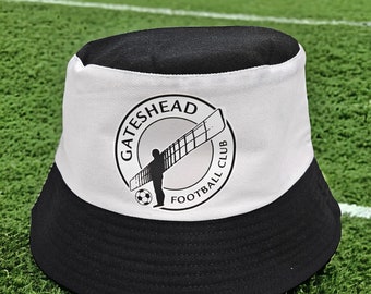 Puede incluir: Un gorro de cubo negro y blanco con un logotipo blanco que dice "Gateshead Football Club" y un gráfico de una persona sosteniendo un ala grande.