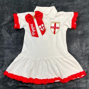 May include: White polo dress with red trim and a red bow with the name "Pippa". The dress has a collared neckline, short sleeves with red frills, and a ruffled hem. An embroidered emblem with the text "IT'S COMING HOME" and "WORLD CUP '66" is on the chest.