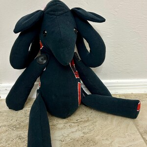 Puede incluir: Un juguete de peluche negro hecho a mano con orejas y extremidades largas. El juguete tiene ojos de botón y está acentuado con tela roja y blanca. Los pies tienen círculos blancos y detalles en rojo. El juguete está sentado en un suelo embaldosado.