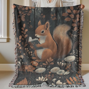 May include: A woven tapestry throw blanket features a whimsical illustration of a squirrel reading a book in a forest setting. The squirrel is brown and orange with a bushy tail. The background is a dark forest with green leaves and flowers.