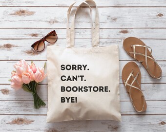 May include: A white canvas tote bag with black text that reads "SORRY. CAN'T. BOOKSTORE. BYE!" The bag is on a white wooden surface with a bouquet of pink tulips, sunglasses, and two pairs of brown sandals.