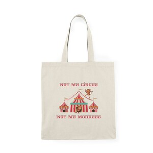 Peut inclure: Un sac fourre-tout beige avec le texte "Not my circus, not my monkeys" imprimé en rouge. Le design présente une tente de cirque rouge et blanche à rayures avec des singes suspendus au sommet.