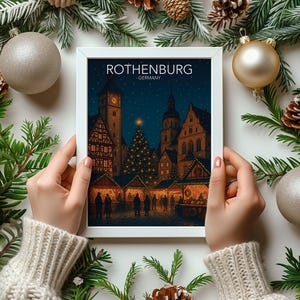 May include: A framed print of Rothenburg, Germany, held by hands, surrounded by Christmas decorations and fir branches. The print shows a night scene of a Christmas market with lit stalls and a decorated tree. The text "ROTHENBURG GERMANY" is at the top.