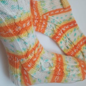 Könnte beinhalten: Ein Paar handgestrickte Socken in einem farbenfrohen Muster. Die Socken haben weiße, orangefarbene, gelbe und türkisfarbene Akzente. Das Design umfasst Zopfstrickdetails und gestreifte Abschnitte. Die Socken sind aus einem weichen, strukturierten Garn gefertigt.
