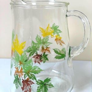 May include: Clear glass pitcher with a handle and a lid. The pitcher is decorated with colorful maple leaves in shades of green, yellow, and brown. The pitcher is designed for serving beverages.