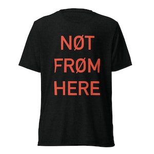 May include: A black short-sleeved t-shirt featuring the text NØT FRØM HERE printed in a bold, coral-orange colour across the chest. The shirt has a crew neckline and a standard fit, with the letter O replaced by the slashed zero symbol.