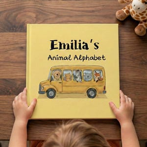 May include: A yellow children's book titled "Emilia's Animal Alphabet" featuring a school bus illustration with various animals. The book is held by a child on a wooden surface, with a giraffe plush toy in the background.