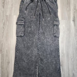 May include: Stone-washed gray cargo pants with a wide leg design. The pants feature two cargo pockets on the sides, a drawstring waist, and a relaxed fit. The fabric has a textured appearance.