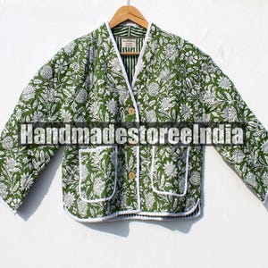 以下が含まれることがあります： 2つのフロントポケットと木製ボタンが付いた、緑と白の花柄プリントのジャケット。 ジャケットは白いトリムで、木製のハンガーに吊るされています。 「handmadestoreindia」の文字が前面に印刷されています。