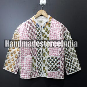 May include: A quilted jacket with a floral pattern in white, pink, and green. The jacket has a zip closure, two pockets, and is hanging on a wooden hanger. The text "HandmadestoreIndia" is across the front.