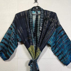 May include: A tie-dye kimono robe in shades of blue, black, and yellow hangs from a black hanger. The robe features a tie-dye pattern with horizontal stripes on the sleeves and a knotted belt. The background is a white brick wall.
