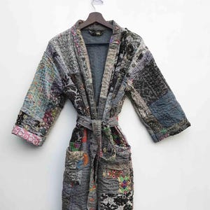 May include: A patchwork robe in shades of gray, blue, and black, with a tie belt. The robe features a variety of patterned fabric squares, creating a unique, textured design. The sleeves are three-quarter length, and the robe is hanging on a wooden hanger.