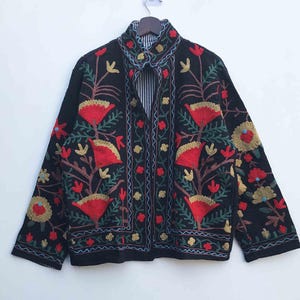 Peut inclure: Veste noire brodée de motifs floraux rouges, jaunes et verts. La veste a un col et des manches longues. La broderie comprend des fleurs et des feuilles stylisées, créant un motif décoratif. La veste est suspendue sur un fond blanc.