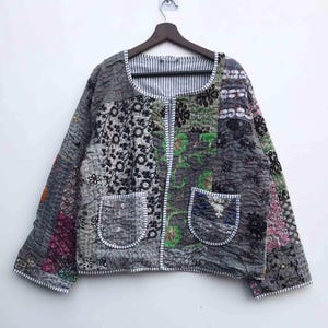 Könnte beinhalten: Graue Patchwork-Jacke mit verschiedenen floralen und geometrischen Mustern. Sie hat zwei Vordertaschen, ein gestreiftes Futter und einen Reißverschluss. Die Jacke hängt an einem Holzbügel.
