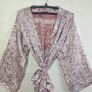 May include: A floral patterned robe in shades of white and red, hanging on a black hanger. The robe has wide sleeves and a tie closure at the waist. The fabric appears to be silky and lightweight, with a detailed floral design.