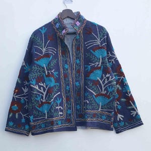 May include: A blue jacket with intricate floral embroidery in teal, burgundy, and white. The jacket features a high collar and a striped lining. The embroidery design includes stylized flowers and vines, creating a detailed and artistic look.