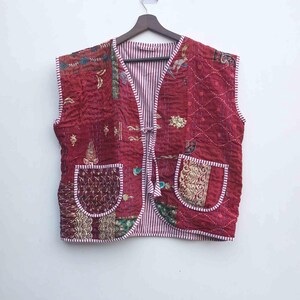 May include: A sleeveless, quilted vest in shades of burgundy, with patterned patches and two front pockets. The vest has a striped lining and is trimmed with white piping. It hangs from a wooden hanger against a white background.