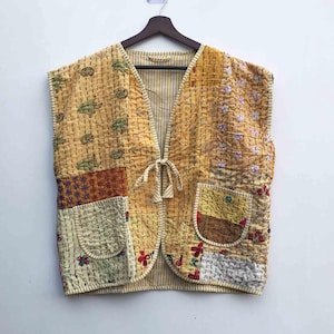 Peut inclure: Un gilet sans manches en patchwork dans les tons de jaune, beige et marron. Le gilet présente divers motifs de tissu, dont des motifs floraux et géométriques, avec une fermeture à nouer à l'avant. Il est suspendu à un cintre en bois sur un mur blanc.
