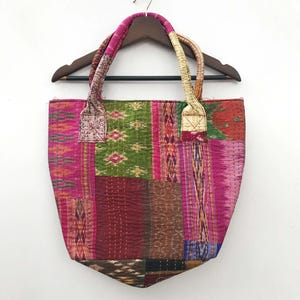 Könnte beinhalten: Eine Patchwork-Tragetasche in verschiedenen Farben, darunter Rosa, Grün und Burgunderrot. Die Tasche hat eine rechteckige Form mit abgerundetem Boden und zwei Griffen aus gedrehtem Stoff. Sie hängt an einem Holzkleiderbügel. Die Tasche besteht aus verschiedenen Stoffflicken.