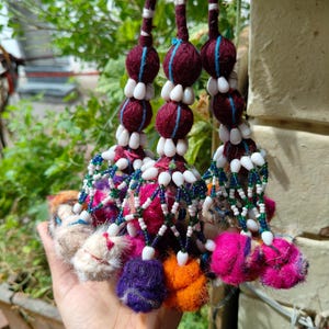 Könnte beinhalten: Ein dekorativer Hängeschmuck mit drei vertikalen Strängen, die mit kastanienbraunem und gelbem Garn umwickelt sind. Jeder Strang enthält kastanienbraune Stoffkugeln, weiße Kaurimuschel-Perlen und ein Netz aus grünen und weißen Perlen mit flauschigen Bommeln.