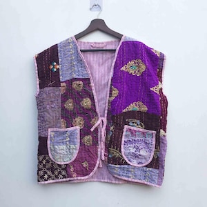 以下が含まれることがあります： 紫、ラベンダー、バーガンディの色合いのパッチワークベスト。刺繍のディテールと2つのフロントポケットが付いています。ベストはストライプの裏地とタイクロージャーが付いています。この衣服はノースリーブで、白い壁に木製のハンガーに掛けられています。