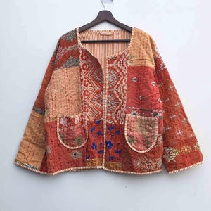 Pode incluir: Casaco patchwork em tons de vermelho, laranja e bege, com detalhes bordados intrincados e dois bolsos frontais. O casaco é feito de vários padrões e texturas de tecido e está pendurado em um cabide de madeira.