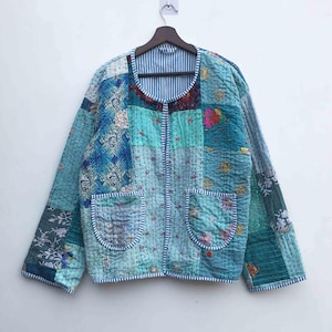 May include: A patchwork jacket in shades of blue, teal, and green, with floral and geometric patterns. The jacket has two front pockets and a striped collar. The jacket is hanging on a wooden hanger.