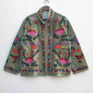 Peut inclure: Une veste vert sauge avec des broderies florales vibrantes. La veste a un col montant et une fermeture éclair sur toute la longueur. La broderie comprend des fleurs roses, rouges, oranges et bleues avec des tiges et des feuilles noires. La veste est suspendue à un cintre en bois.