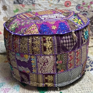 Op de afbeelding: Een ronde patchwork poef in tinten paars, goud en blauw. De boven- en zijkanten zijn versierd met ingewikkelde geborduurde patronen en een witte kralenrand. Een decoratief accentstuk.