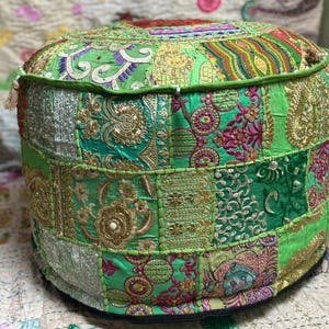May include: A round, patchwork pouf in shades of green, gold, and silver. The fabric is intricately embroidered with floral and geometric patterns. The pouf has a black base and a decorative tassel.