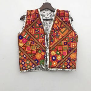 Può includere: Un gilet ricamato con un design patchwork. Il gilet presenta un vivace mix di motivi geometrici e floreali rossi, arancioni, rosa e verdi. Ha una fodera bianca a fantasia floreale ed è appeso a una gruccia di legno.