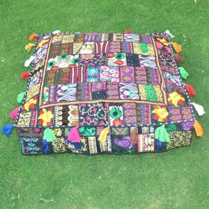 Könnte beinhalten: Quadratisches Patchwork-Kissen mit einer lebendigen Mischung aus Farben und Mustern. Das Kissen weist eine Vielzahl von bestickten Quadraten in Rot-, Blau-, Grün- und Goldtönen auf. Bunte Quasten säumen die Ränder und verleihen eine dekorative Note.