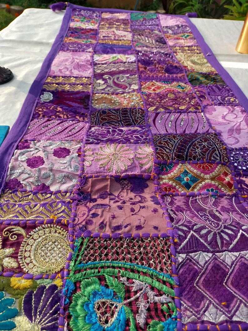 Peut inclure: Chemin de table patchwork violet, compos&eacute; de divers tissus et textures. Le chemin de table rectangulaire a une bordure violette unie et est orn&eacute; de motifs floraux et g&eacute;om&eacute;triques brod&eacute;s. Mesure environ 150 cm de long.