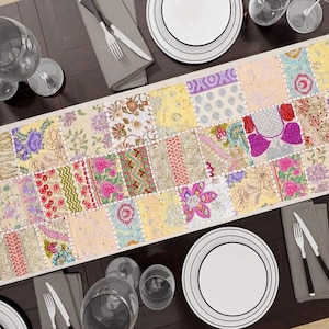以下が含まれることがあります： 様々な花柄と幾何学模様がパッチワークされたカラフルなテーブルランナー。濃い色の木製テーブルの上に置かれ、白いお皿、カトラリー、ワイングラスに囲まれています。ランナーのデザインには、ピンク、黄色、紫の色合いが含まれています。